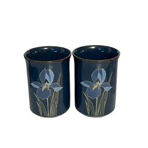 May include: Two dark blue ceramic mugs with a light blue iris flower design. The mugs have a gold rim and a cylindrical shape. The iris flower has light blue petals and a yellow center with thin, light beige stems.