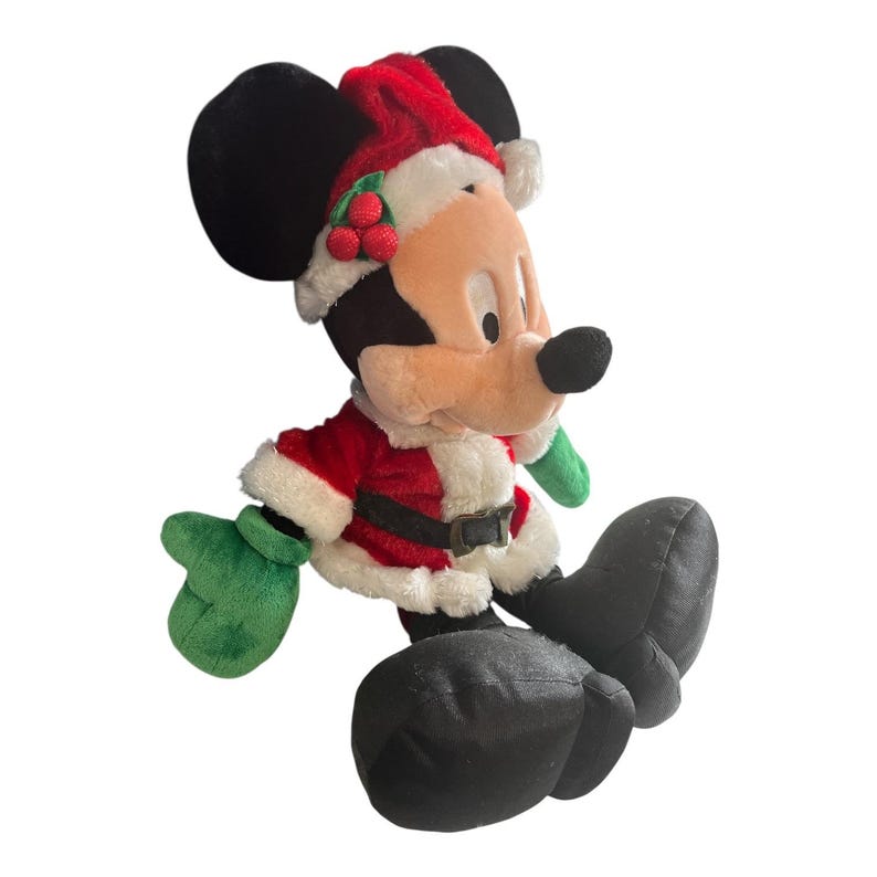 Puede incluir: Un peluche de Mickey Mouse vestido con un traje de Pap&aacute; Noel. La chaqueta roja tiene ribetes blancos y un cintur&oacute;n negro. Lleva un gorro de Pap&aacute; Noel con bayas de acebo y guantes verdes. Sus orejas y zapatos negros son visibles.