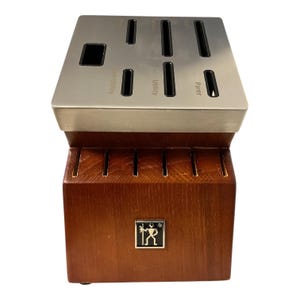 May include: A wooden knife block with a stainless steel top. The top has slots labeled for different knife types: serrated utility, utility, and parer. A logo is on the front of the block.