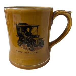 Puede incluir: Una taza de cerámica vintage con un esmalte marrón y un borde dorado. La taza presenta una ilustración en blanco y negro de un coche antiguo con el texto "WHITE STEAM CAR". El asa es curva y la taza tiene una forma ligeramente acampanada.