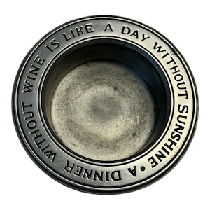 May include: A circular, pewter-colored wine coaster with the engraved text "A dinner without wine is like a day without sunshine." The coaster has a slightly textured surface and a raised rim.