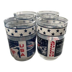 May include: Set of four clear glass tumblers. Each tumbler features a white band with blue stars, and a design with the text "USA" and "APOLLO 13". The glasses have a vintage aesthetic and are likely collectible.