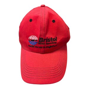 May include: A bright red baseball cap featuring black stitching and black eyelets. The front displays an embroidered logo for Bristol Motor Speedway with the text "Bristol Motor Speedway" and the slogan "Racin' the way it oughta be".