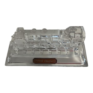 May include: A clear glass train ornament with the words "Orient Express" on a small rectangular plaque. The train is detailed with various components, including a smokestack and wheels, and sits on a rectangular base.