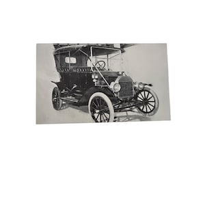 May include: A black and white photograph of a vintage automobile with a soft top, large spoked wheels, and a prominent front radiator grille. The vehicle features a boxy body design, a steering wheel visible through the windshield, and round headlights.