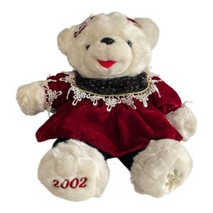 May include: A white plush teddy bear wearing a red velvet dress with white lace trim and a black collar. The bear has a red smiling mouth and the year "2002" embroidered on one paw. A gold snowflake is embroidered on the other paw.