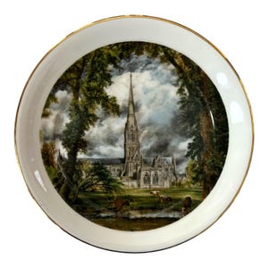 May include: A round, white decorative plate with a gold rim. The plate features a detailed painting of a cathedral framed by trees, with cows grazing in a field. The sky is overcast with dark clouds.