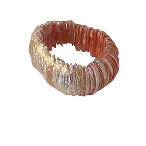 Puede incluir: Un brazalete nacarado con tonos coral e iridiscentes. Está compuesto por pequeños elementos con forma de concha, creando un accesorio texturizado y llamativo. El diseño es circular y parece ser elástico, adaptable a diferentes muñecas.
