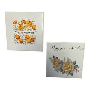 May include: Two white ceramic tiles. One tile features the word "CALIFORNIA" with orange flowers and oranges. The other tile has the text "Peggy's Kitchen" with yellow and pink roses.
