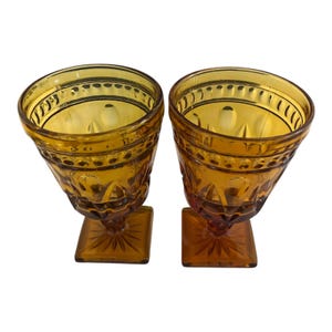 May include: Two amber-colored vintage goblets. Each glass features a square base, a detailed stem, and a patterned bowl. The glasses are made of a transparent material, allowing light to pass through. The goblets are ideal for serving beverages.