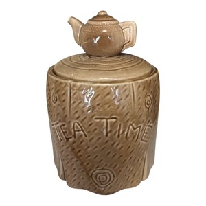 May include: A brown ceramic tea time jar with a teapot-shaped lid. The jar has a textured, tree-trunk-like design with the words "TEA TIME" carved into the front. Decorative swirls and circles are also carved into the jar.