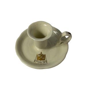 May include: Cream-colored ceramic candlestick holder with a circular base and a small handle. The base features a gold emblem with text that reads "Seaview Country Club, Absecon, New Jersey."