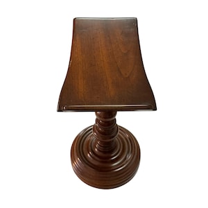 May include: A dark brown wooden book stand featuring a curved, rectangular top surface with a small ledge at the bottom. The stand is supported by a central, turned pedestal column that rests on a circular, tiered base with concentric rings.