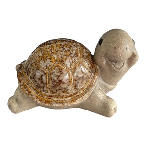 Może przedstawiać: Ceramiczna figurka żółwia z uśmiechniętą twarzą. Skorupa żółwia to mozaika brązu, beżu i bieli, a głowa i kończyny są jasnoszare. Figurka jest przedmiotem dekoracyjnym.