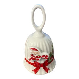 Puede incluir: Una campana de cerámica blanca con textura acanalada vertical, que presenta una figura en relieve de Santa Claus sosteniendo un bastón de caramelo. La base está decorada con un lazo rojo y hojas de acebo verdes con bayas rojas pintadas.