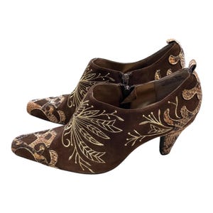 May include: A pair of brown suede ankle boots with a pointed toe and a small heel. The boots feature gold embroidered leaf designs and faux snakeskin accents. A zipper runs up the side of each boot.
