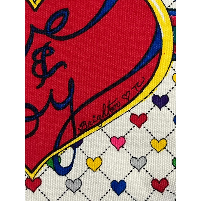 Op de afbeelding: Close-up van een stof met een groot rood hartontwerp en de woorden "Love & Joy" in blauw schrift. Het hart is geel en blauw omlijnd. De achtergrond is wit met een patroon van kleine harten in verschillende kleuren.