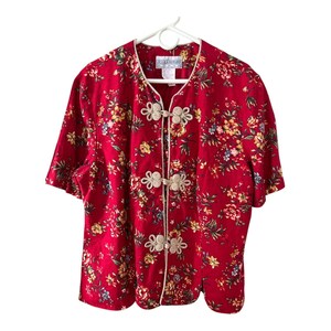 May include: A red floral print blouse with short sleeves and decorative frog closures. The blouse features a white trim and a vibrant pattern of yellow, blue, and green flowers. The tag reads "Jesse Howard".