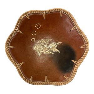 May include: A brown decorative bowl with a woven rim. The bowl features a fish design in the center, with white dots and a black gradient on the right side. The rim is made of woven material and is attached to the bowl with small, light-colored pieces.
