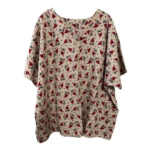 May include: A beige, short-sleeved top with a V-neck and two pockets. The top is covered in a repeating pattern of red Santa hats and green sprigs, creating a festive holiday design. The fabric appears to be a lightweight material.