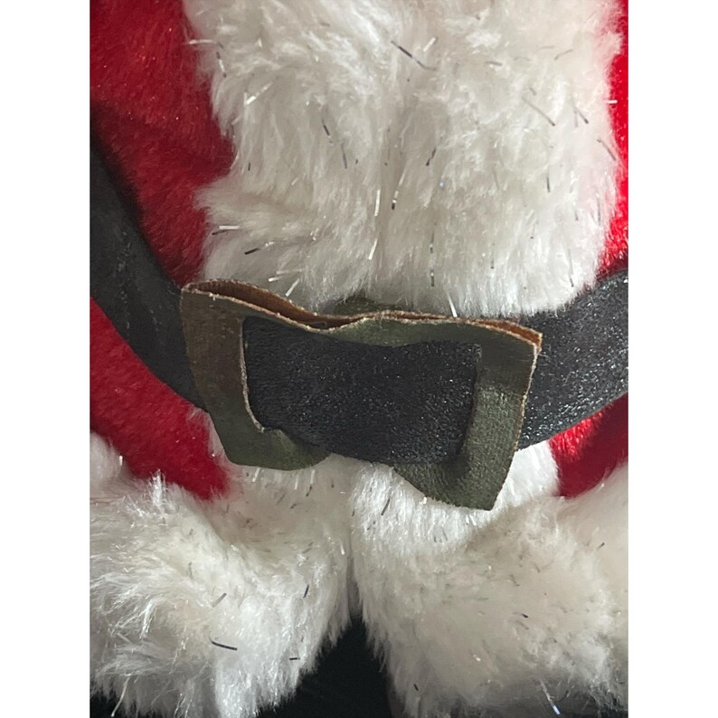 Puede incluir: Primer plano de un mu&ntilde;eco de peluche de Pap&aacute; Noel. El mu&ntilde;eco presenta un abrigo rojo, ribete de piel sint&eacute;tica blanca y un cintur&oacute;n negro con hebilla dorada. La piel blanca tiene detalles de oropel plateado.