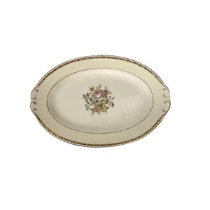 Puede incluir: Una bandeja de servir ovalada de color crema con un ramo de flores central en tonos rosa, amarillo y verde. El borde está decorado con un patrón de filigrana en tono dorado e incluye asas integradas con detalles en forma de voluta.