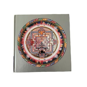 May include: A square book with a silver cover featuring a large, circular, multi-colored mandala illustration. The intricate design includes geometric patterns, small figures, and symbols in shades of red, blue, orange, and white, centered on the front.