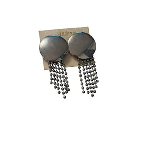Peut inclure: Une paire de boucles d'oreilles circulaires argentées fixées sur un carton de présentation beige portant l'inscription Andante. Chaque boucle présente un disque lisse et poli surmontant plusieurs chaînes pendantes de petites perles métalliques.