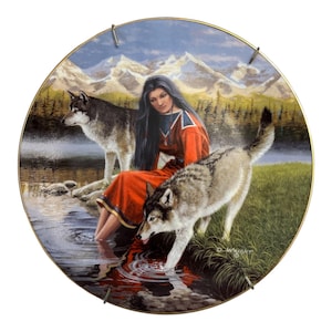Puede incluir: Plato decorativo redondo que representa a una mujer nativa americana con un vestido rojo con dos lobos cerca del agua. El fondo incluye montañas, árboles y un cielo azul. El plato tiene un borde dorado y herrajes metálicos para colgar.