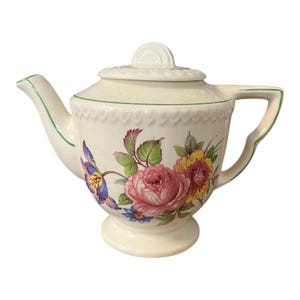May include: Cream-colored ceramic teapot with a floral design featuring pink roses, yellow flowers, and blue accents. The teapot has a green trim, a lid, and a handle. The teapot is a vintage style.