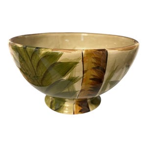 May include: A ceramic bowl with a pedestal base, featuring a hand-painted design of green palm leaves and brown tree trunks against a beige background. The bowl has a wide, open top and a glossy finish.