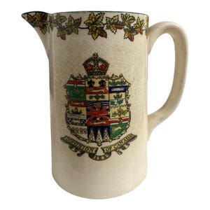 May include: Cream-colored ceramic pitcher with a handle and spout. The pitcher features a detailed coat of arms with a crown and the words "Dominion of Canada." The rim is decorated with a garland of leaves.