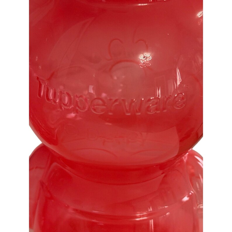 May include: A translucent red plastic container with a rounded shape and a ribbed base. The word Tupperware is embossed in the center of the surface. The material is smooth and glossy, reflecting light across its curved, semi-transparent body.