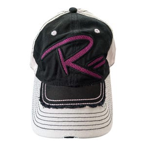 May include: A baseball cap featuring a black front panel with a large, embroidered magenta letter R. The cap has a distressed, frayed brim with light gray fabric and black stitching. The side panels are light gray with matching black stitching.