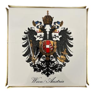 May include: Square white decorative tile with a gold trim. The tile features a detailed black and gold double-headed eagle emblem with a crown, shield, and sword. The text "Wien / Austria" is printed at the bottom.
