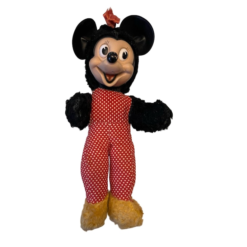 May include: Vintage Mickey Mouse plush doll. The doll has a black head and ears, a red and white polka dot jumpsuit, and tan shoes. A red bow is on top of the head. The doll is standing with its arms out.