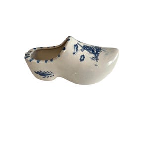 May include: A ceramic Dutch clog-shaped planter. The white planter features blue windmill and floral designs. The rim and heel of the clog are also decorated with blue accents. The planter is a decorative item.