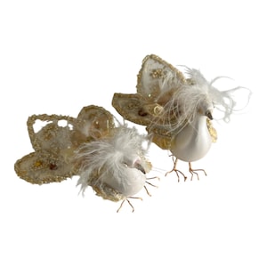 Può includere: Due figurine decorative di uccelli. Gli uccelli hanno corpi bianchi, ali dorate e soffici accenti di piume bianche. Le ali sono impreziosite da perline e paillettes dorate. Gli uccelli hanno delicate zampe di filo su sfondo bianco.