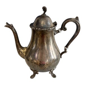 Puede incluir: Una tetera plateada con un pico y asa curvados. La tetera tiene una tapa con un adorno decorativo y se apoya sobre tres pequeños pies. El diseño es ornamentado y vintage, con una superficie reflectante.