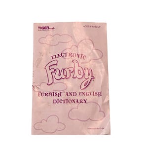May include: A pink Furby Furbish and English Dictionary. The cover features the word "Furby" in a cursive font, with the words "Electronic" above and "Furbish and English Dictionary" below. The cover also has cloud illustrations.