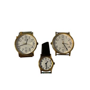 May include: Three vintage Timex watches with gold-toned cases and white faces. The watches have black hands and Arabic numeral hour markers. One watch has a black leather band, while the others have metal bands. The Timex logo is visible on the watch faces.