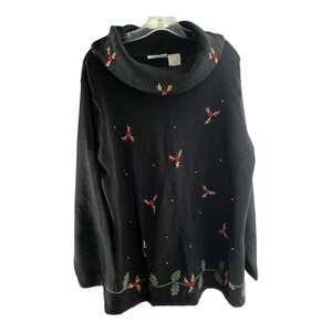 May include: Black sweater with a cowl neck and long sleeves. The sweater is decorated with embroidered red berries and green leaves, with white dots scattered across the front. The bottom hem and neckline are trimmed with red stitching.