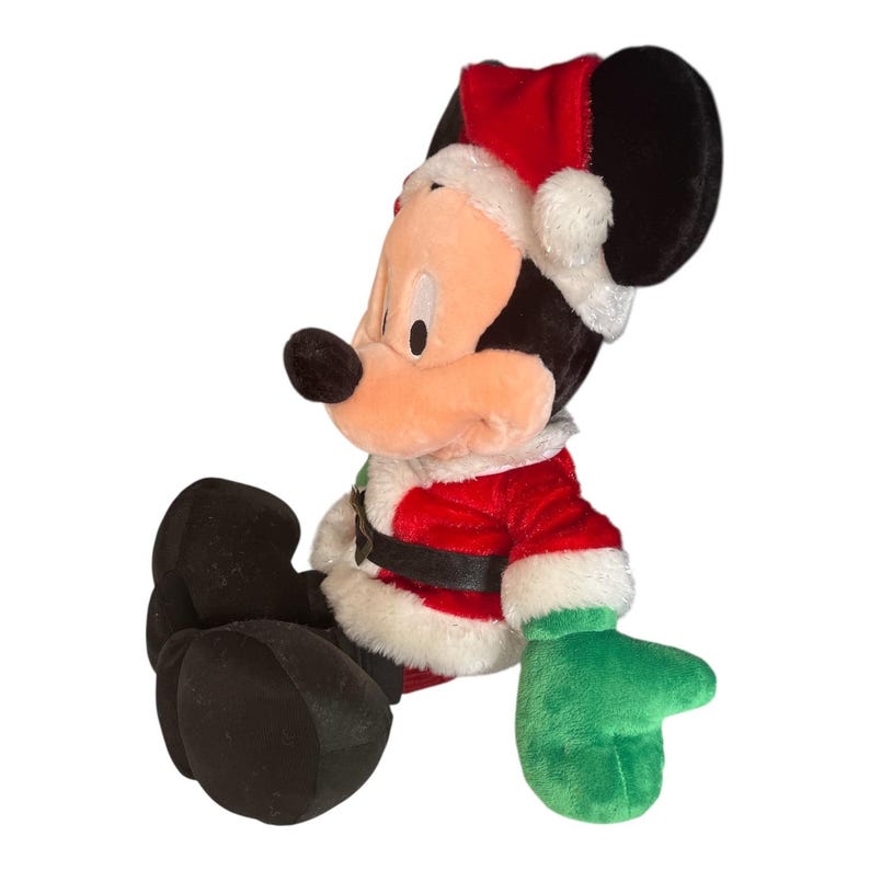 Puede incluir: Un peluche de Mickey Mouse vestido con un traje de Pap&aacute; Noel. La chaqueta roja tiene ribetes blancos y un cintur&oacute;n negro. Mickey lleva un gorro de Pap&aacute; Noel y guantes verdes. El peluche est&aacute; sentado.