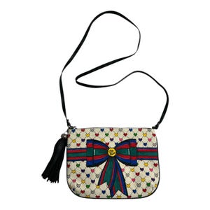 Op de afbeelding: Cr&egrave;mekleurige crossbodytas met een zwarte band en kwast. De tas heeft een patroon van kleurrijke harten en een groot strikdesign met rode, groene en blauwe strepen. De strik heeft een goudkleurig embleem in het midden.
