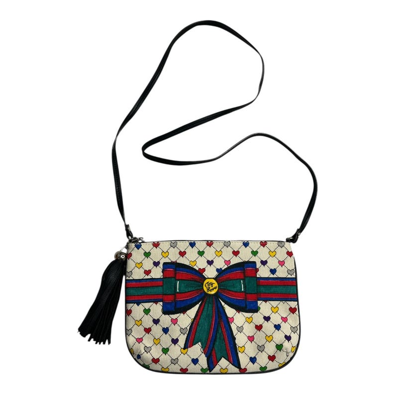 Op de afbeelding: Cr&egrave;mekleurige crossbodytas met een zwarte band en kwast. De tas heeft een patroon van kleurrijke harten en een groot strikdesign met rode, groene en blauwe strepen. De strik heeft een goudkleurig embleem in het midden.