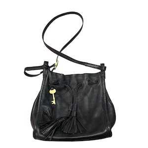 Könnte beinhalten: Eine schwarze Beuteltasche aus genarbtem Leder mit Kordelzugverschluss, zwei schwarzen Lederquasten und einem goldfarbenen Metallschlüsselanhänger an einem dünnen Riemen. Die Tasche verfügt über einen verstellbaren Schulterriemen mit goldfarbenen Beschlägen.