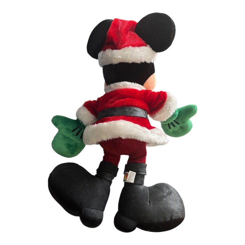 Puede incluir: Un peluche de Mickey Mouse vestido con un traje de Pap&aacute; Noel. La figura lleva una chaqueta roja con ribete blanco, un cintur&oacute;n negro y un gorro de Pap&aacute; Noel a juego. Tiene guantes verdes y zapatos negros grandes. La figura est&aacute; de espaldas a la c&aacute;mara.