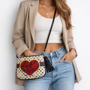 Op de afbeelding: Een crossbodytas met een cr&egrave;mekleurige achtergrond bedekt met kleine kleurrijke hartjes. De tas heeft een groot rood hart met de tekst "Love & Joy". De tas heeft een zwarte band en een zwarte kwast.