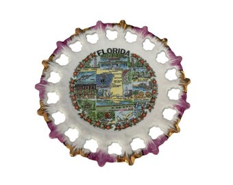 May include: A round ceramic souvenir plate featuring a map of Florida with various illustrations of landmarks and wildlife. The plate has a white center, a scalloped openwork border with purple and gold trim, and the word FLORIDA at the top.