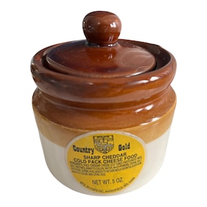 May include: A small, vintage ceramic crock with a lid. The crock is two-toned, with a white base and a brown upper section and lid. A yellow label reads "Country Gold Sharp Cheddar Cold Pack Cheese Food".